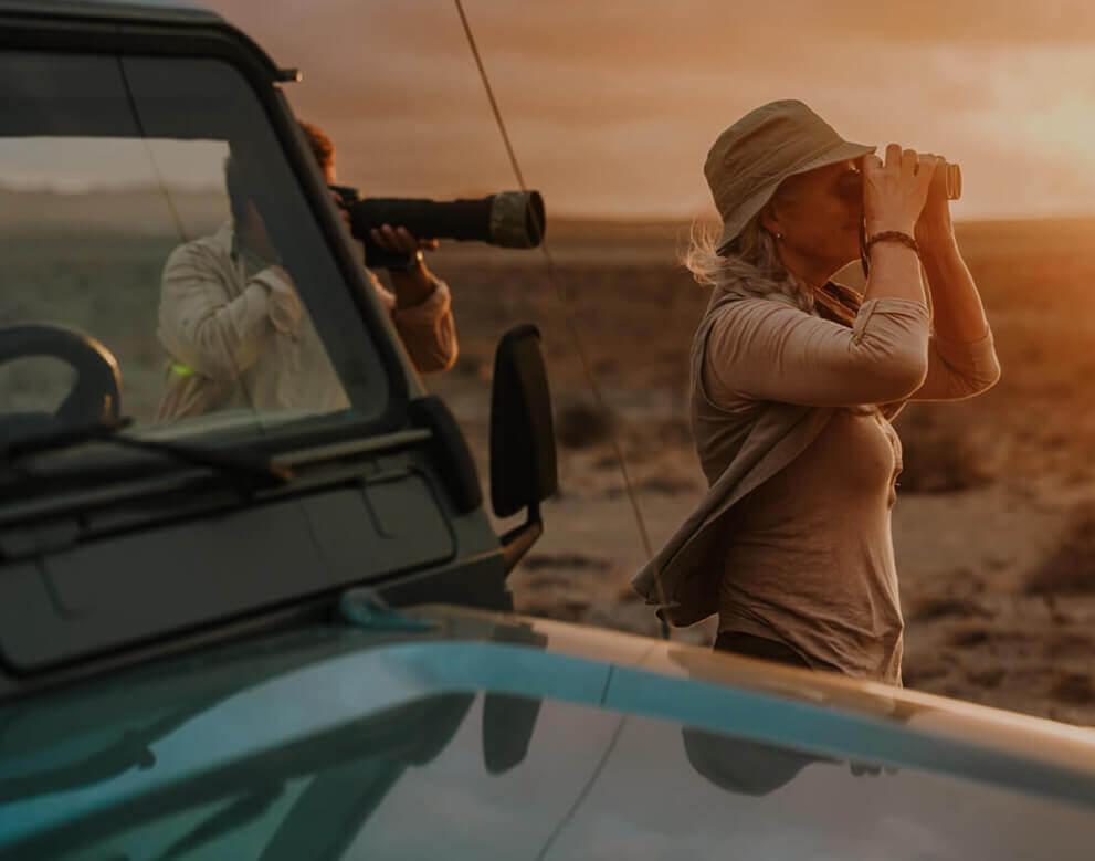 En person bredvid ett fordon i ett kargt landskap observerar fåglar med en kikare i solnedgången.