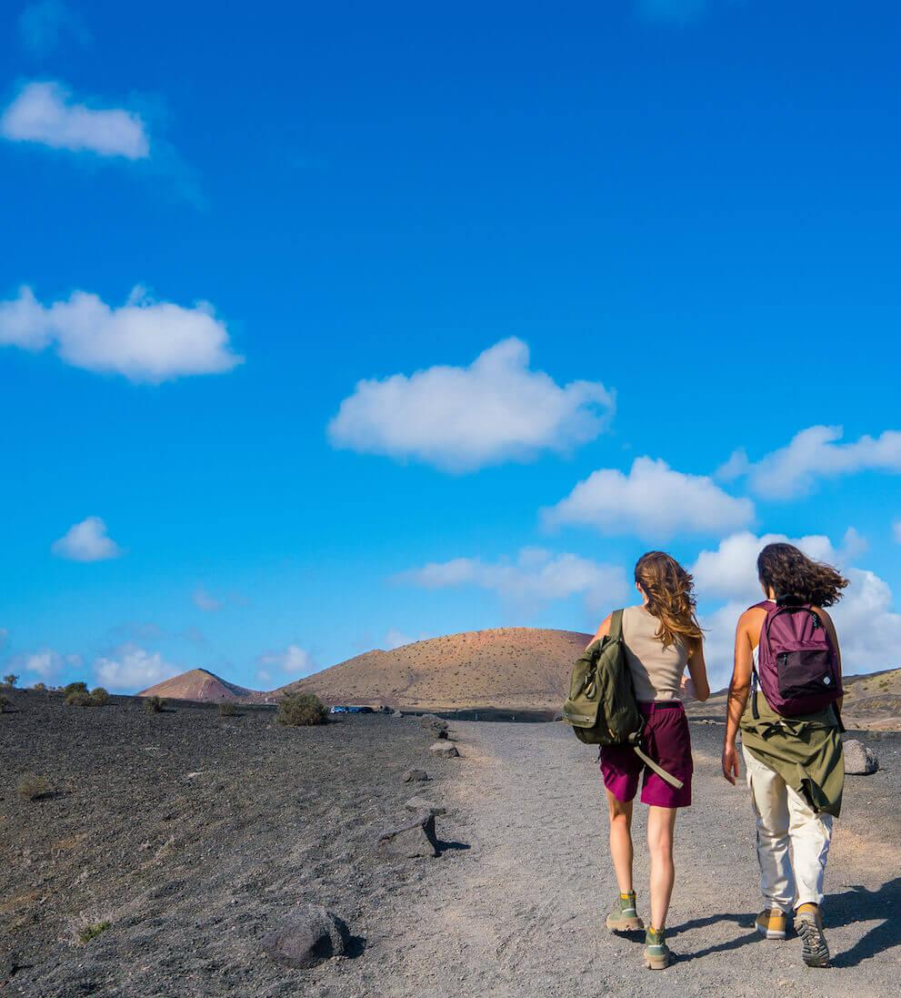 Två personer vandrar under blå himmel längs en vulkanisk vandringsled i Timanfaya Nationalpark.